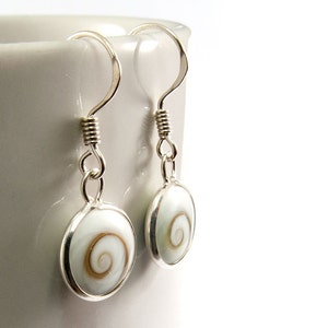 May include: A pair of silver earrings with white oval pendants. Each pendant features a brown spiral design. The earrings are suspended from silver hooks with coiled wire detailing, showcasing a minimalist and elegant design.