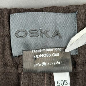 Może przedstawiać: Ciemnobrązowa odzież z szarą metką z nazwą marki OSKA w kolorze czarnym. Mniejsza czarna metka poniżej zawiera tekst Hose Alana long, UDHO36 C08 oraz info@oska.de, a biała metka wskazuje liczbę 505.