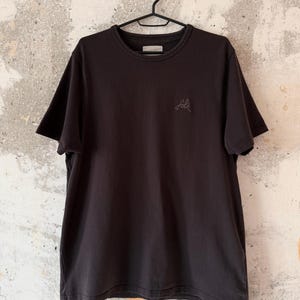 May include: A black crew neck t-shirt hanging on a black hanger. The shirt has short sleeves and a small, embroidered logo on the chest. The background is a textured, off-white wall.
