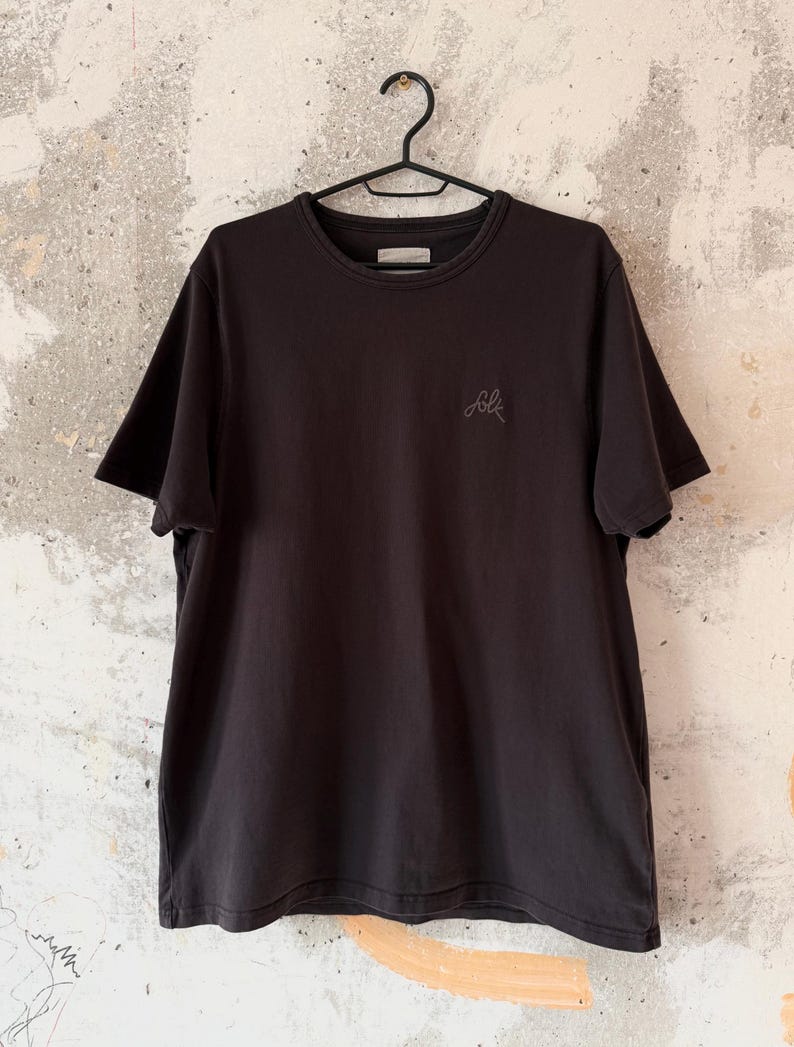May include: A black crew neck t-shirt hanging on a black hanger. The shirt has short sleeves and a small, embroidered logo on the chest. The background is a textured, off-white wall.