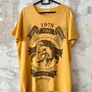 Könnte beinhalten: Senfgelbes T-Shirt mit Rundhalsausschnitt und kurzen Ärmeln. Das Shirt hat eine Grafik mit dem Text "1978 THE BRAVE" und "DIESEL industry" sowie ein Profil einer Person mit Irokesenschnitt.