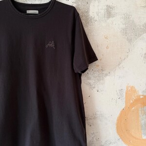 May include: Black crew neck t-shirt with short sleeves, hanging on a black hanger. The word "folk" is embroidered in a light gray script on the front. The shirt is made of a soft material.