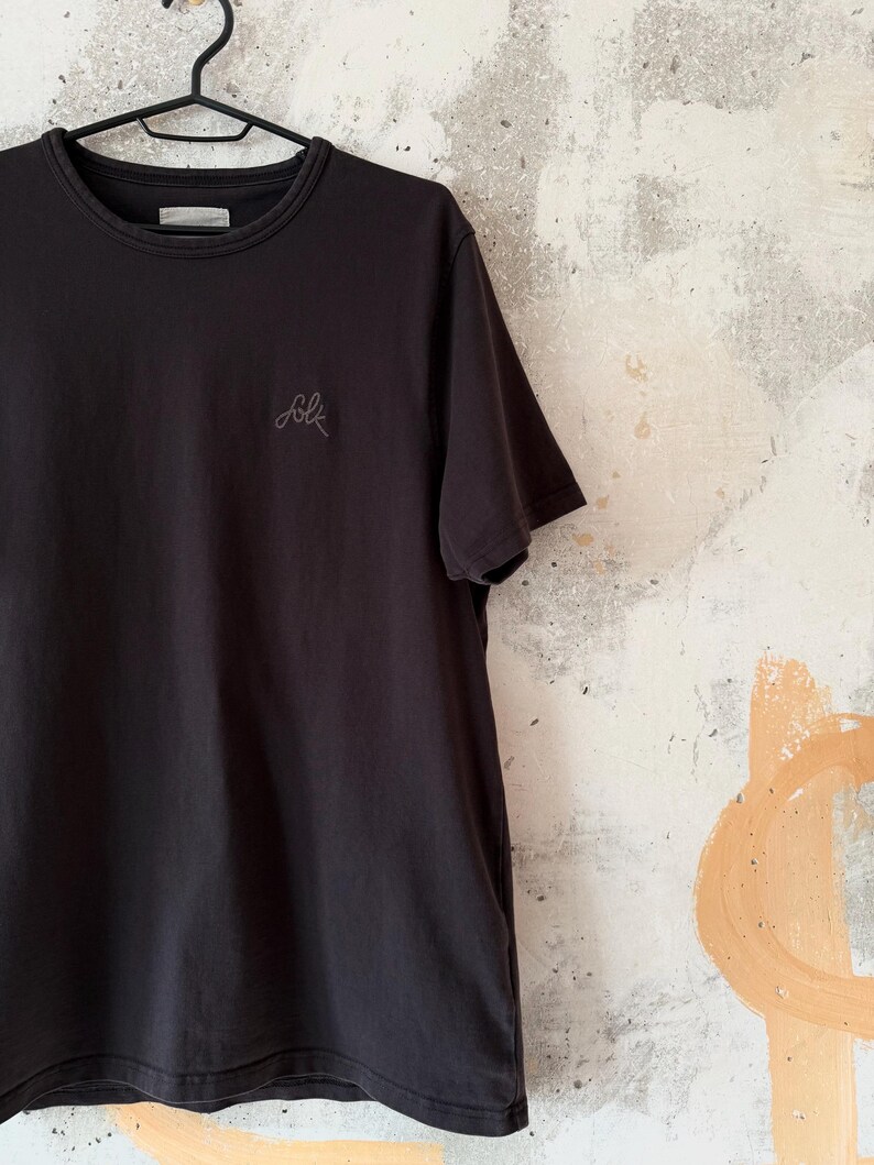May include: Black crew neck t-shirt with short sleeves, hanging on a black hanger. The word "folk" is embroidered in a light gray script on the front. The shirt is made of a soft material.
