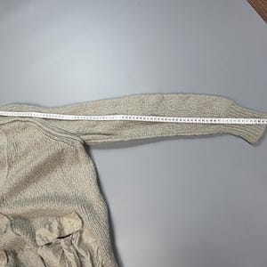 Op de afbeelding: Een beige gebreide trui met een lange mouw en een zakdetail van gladde stof. Een wit meetlint is langs de bovenkant van de mouw geplaatst en toont een lengte van ongeveer 71 cm vanaf de schoudernaad tot aan de rand van de manchet.