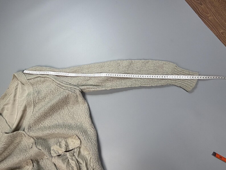 Op de afbeelding: Een beige gebreide trui met een lange mouw en een zakdetail van gladde stof. Een wit meetlint is langs de bovenkant van de mouw geplaatst en toont een lengte van ongeveer 71 cm vanaf de schoudernaad tot aan de rand van de manchet.