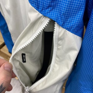 K&ouml;nnte beinhalten: Nahaufnahme einer hellgrauen Jacke mit einem wei&szlig;en Rei&szlig;verschluss. Die Jacke hat ein blau-wei&szlig;es Karomuster am &Auml;rmel und eine Tasche auf der Vorderseite.