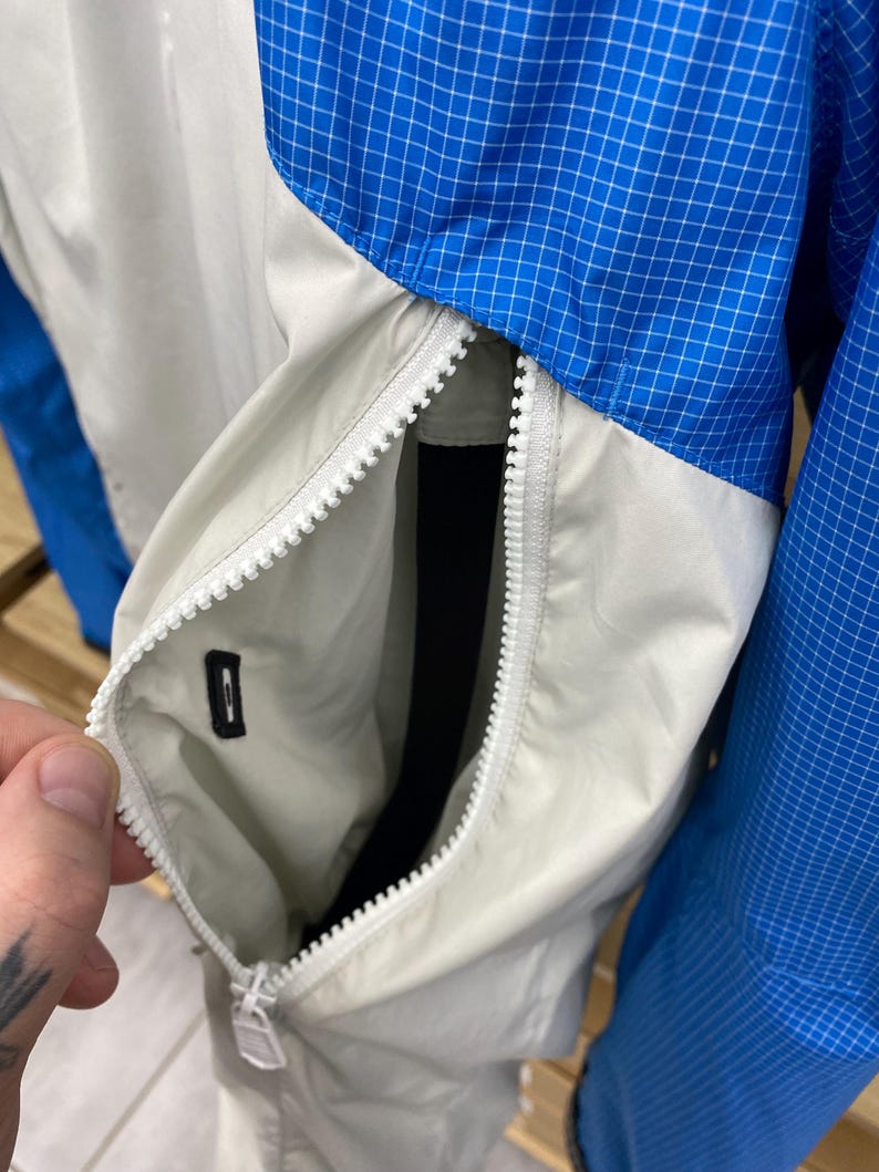 K&ouml;nnte beinhalten: Nahaufnahme einer hellgrauen Jacke mit einem wei&szlig;en Rei&szlig;verschluss. Die Jacke hat ein blau-wei&szlig;es Karomuster am &Auml;rmel und eine Tasche auf der Vorderseite.