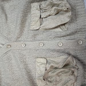 Op de afbeelding: Een beige gebreid vest met een V-hals, een knoopsluiting aan de voorzijde met vijf bijpassende ronde knopen en twee grote, gekreukte, lichtbeige stoffen zakken aan de onderkant. Het kledingstuk is voorzien van een gladde, lichtbeige voering.