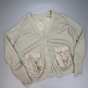 Op de afbeelding: Een beige gebreid vest met een V-hals, lange mouwen met geribbelde boorden en een knoopsluiting aan de voorzijde. Het kledingstuk is voorzien van twee grote, geplooide opgestikte zakken en een gladde, bijpassende beige voering bij de kraag.