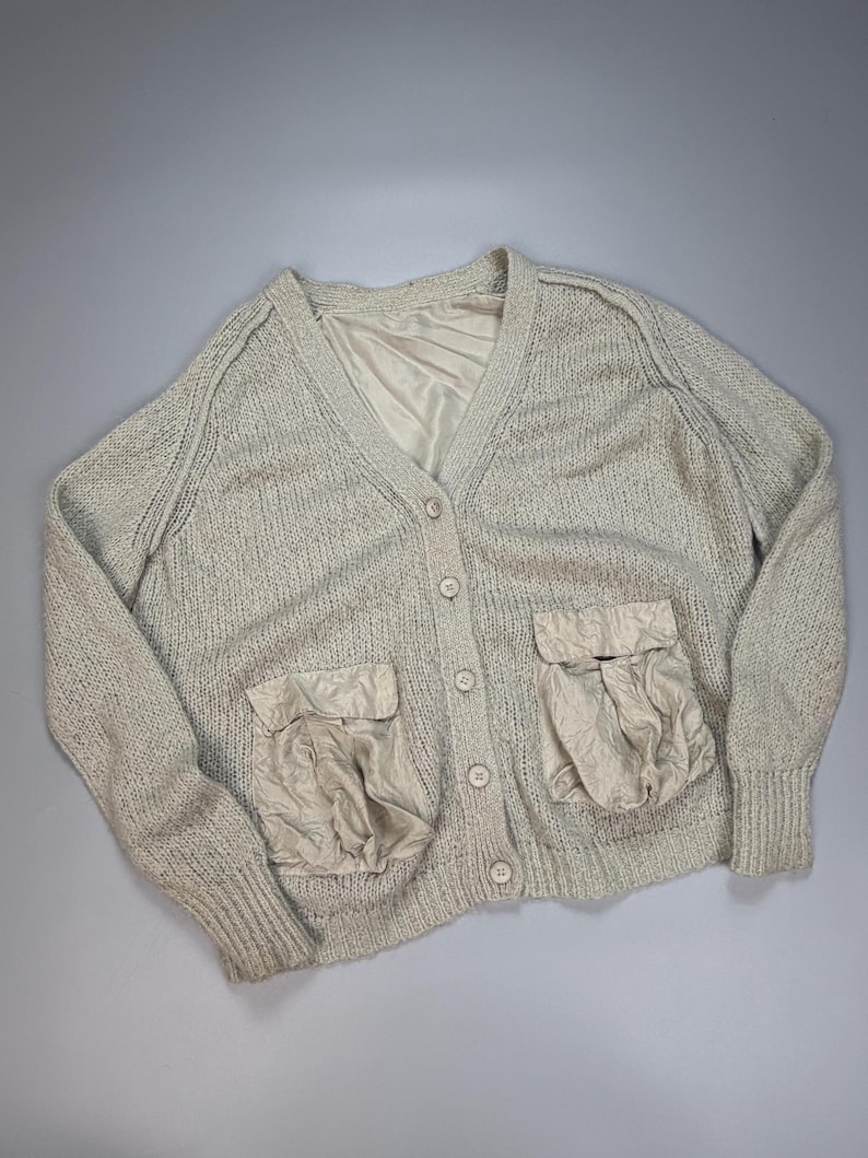 Op de afbeelding: Een beige gebreid vest met een V-hals, lange mouwen met geribbelde boorden en een knoopsluiting aan de voorzijde. Het kledingstuk is voorzien van twee grote, geplooide opgestikte zakken en een gladde, bijpassende beige voering bij de kraag.