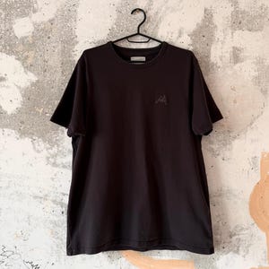 May include: A black crew neck t-shirt hanging on a black hanger. The shirt has short sleeves and a small embroidered design on the chest. The background is a textured, light-colored wall.