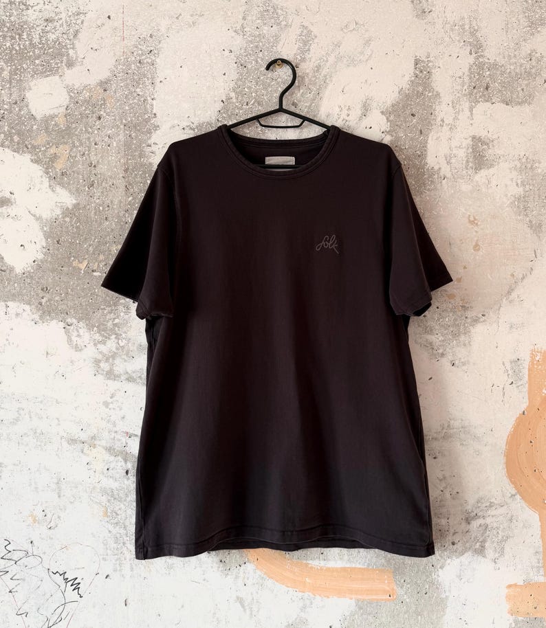 May include: A black crew neck t-shirt hanging on a black hanger. The shirt has short sleeves and a small embroidered design on the chest. The background is a textured, light-colored wall.