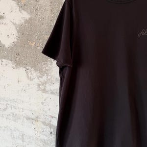 May include: Black short-sleeved t-shirt hanging on a black hanger. The shirt has a crew neck and a small, cursive logo on the chest. The background is a textured, off-white wall.