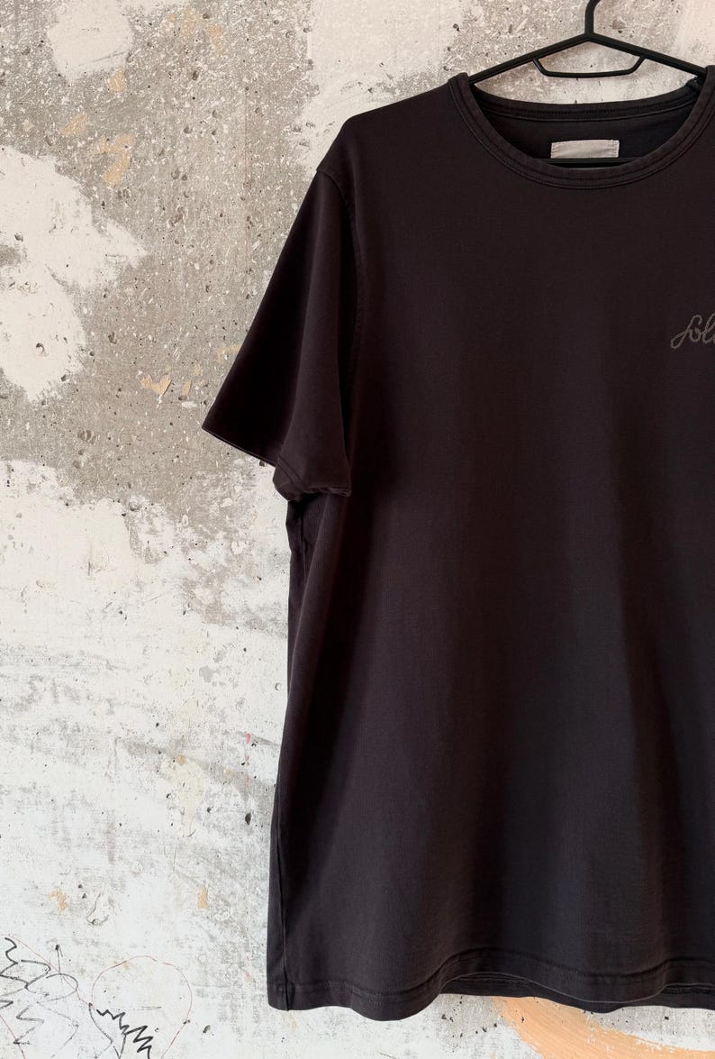 May include: Black short-sleeved t-shirt hanging on a black hanger. The shirt has a crew neck and a small, cursive logo on the chest. The background is a textured, off-white wall.
