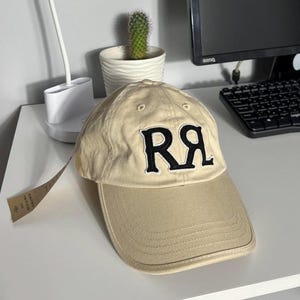 May include: A beige baseball cap featuring a black embroidered RR logo on the front panel. The cap has a curved brim, stitched eyelets, and a brown paper tag attached to the side. The design includes a structured crown and a matching beige strap.
