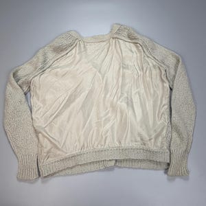 Op de afbeelding: Een trui met lange mouwen met een beige gebreide textuur op de mouwen en rug, gecontrasteerd door een glad, effen beige satijnachtig stoffen paneel aan de voorzijde. Het kledingstuk heeft een geribbelde halslijn en geribbelde boorden aan de mouwen.