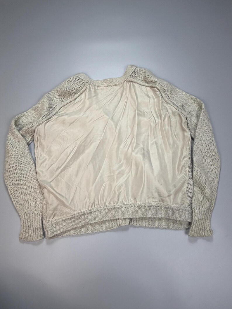 Op de afbeelding: Een trui met lange mouwen met een beige gebreide textuur op de mouwen en rug, gecontrasteerd door een glad, effen beige satijnachtig stoffen paneel aan de voorzijde. Het kledingstuk heeft een geribbelde halslijn en geribbelde boorden aan de mouwen.