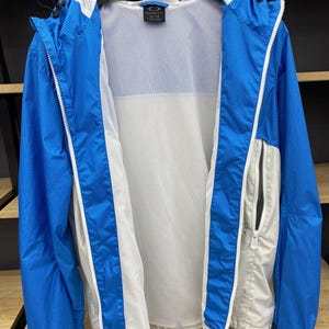 K&ouml;nnte beinhalten: Eine blaue und wei&szlig;e leichte Jacke mit Rei&szlig;verschluss und Mesh-Futter. Die Jacke hat eine Kapuze und eine kleine Tasche auf der linken Seite. Das Etikett auf der Jacke lautet "Oakley Elite".