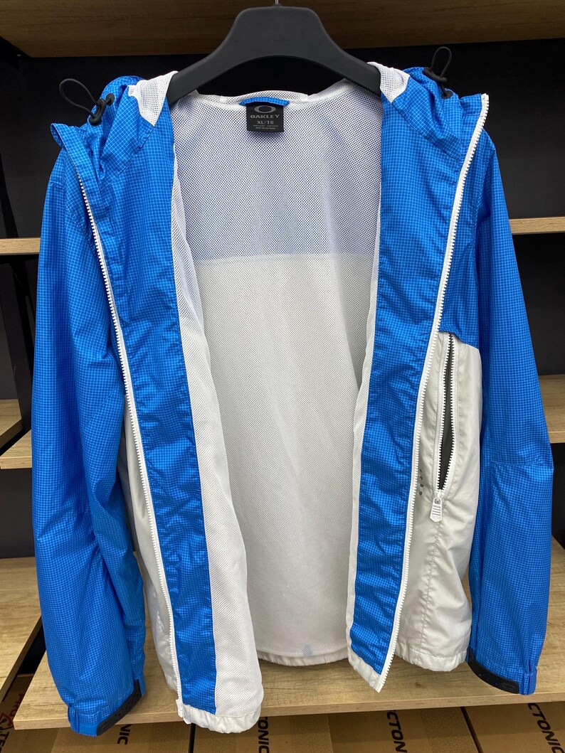 K&ouml;nnte beinhalten: Eine blaue und wei&szlig;e leichte Jacke mit Rei&szlig;verschluss und Mesh-Futter. Die Jacke hat eine Kapuze und eine kleine Tasche auf der linken Seite. Das Etikett auf der Jacke lautet "Oakley Elite".