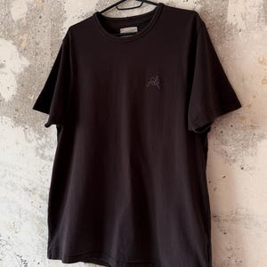 May include: Black crew neck t-shirt hanging on a black hanger. The shirt has short sleeves and a small embroidered "folk" logo on the chest. The shirt is made of a soft material.