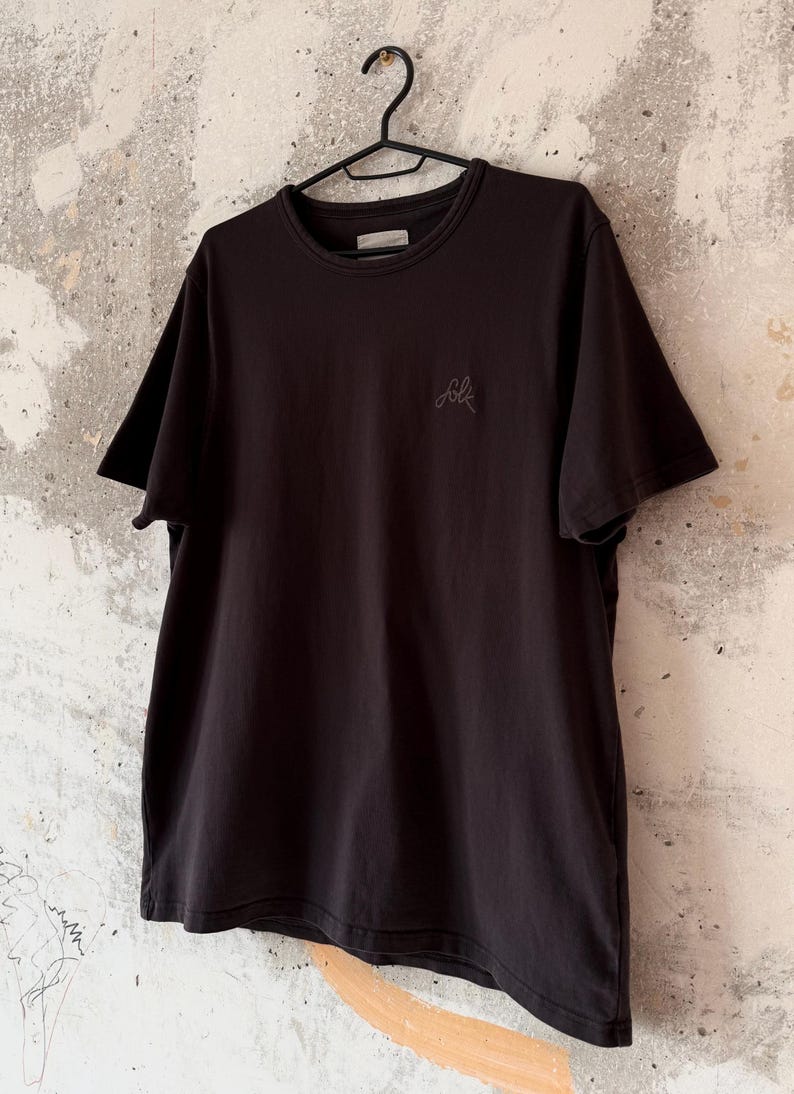 May include: Black crew neck t-shirt hanging on a black hanger. The shirt has short sleeves and a small embroidered "folk" logo on the chest. The shirt is made of a soft material.