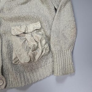 Op de afbeelding: Een lichtbeige gebreid vest met een V-hals, lange mouwen met geribbelde boorden en een enkele knoopsluiting. Het kledingstuk is voorzien van een opvallende, getextureerde opgestikte zak van gekreukte stof aan de voorzijde, wat contrasteert met het breiwerk.