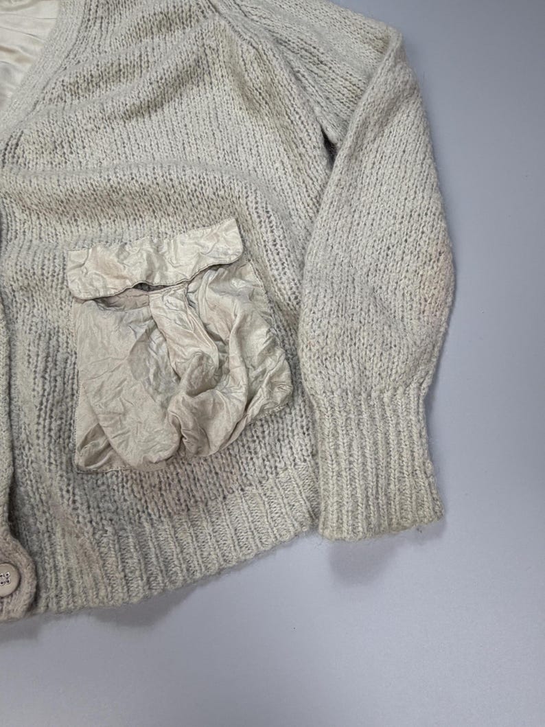 Op de afbeelding: Een lichtbeige gebreid vest met een V-hals, lange mouwen met geribbelde boorden en een enkele knoopsluiting. Het kledingstuk is voorzien van een opvallende, getextureerde opgestikte zak van gekreukte stof aan de voorzijde, wat contrasteert met het breiwerk.