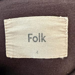 May include: Close-up of a brown fabric with a rectangular label. The label is beige and features the word "Folk" in a dark font. Below the word "Folk" is the number "4". The fabric has a ribbed texture.