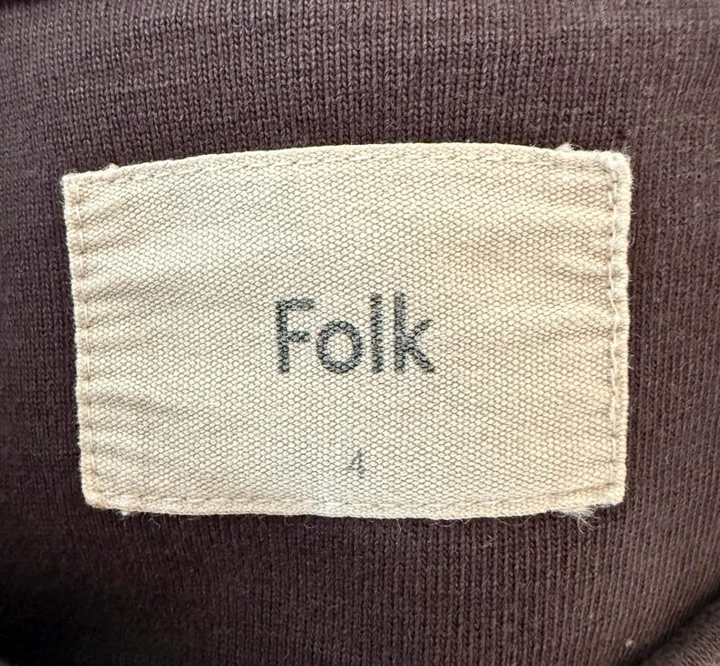 May include: Close-up of a brown fabric with a rectangular label. The label is beige and features the word "Folk" in a dark font. Below the word "Folk" is the number "4". The fabric has a ribbed texture.
