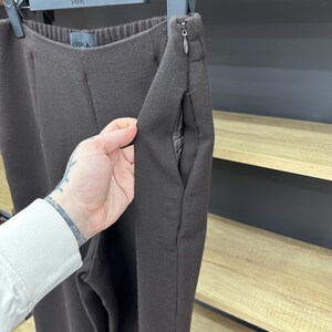 Może przedstawiać: Ciemnobrązowe spodnie z elastycznym pasem, bocznym zapięciem na zamek błyskawiczny z metalowym uchwytem oraz kieszeniami bocznymi. Wnętrze kieszeni wyłożone jest szaro-czarną tkaniną we wzory. Metka marki OSKA jest widoczna w pasie.