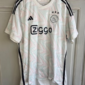 May include: White football shirt with black and red accents. The shirt has a geometric pattern and features the Adidas logo, the Ajax logo, and the text "Ziggo".