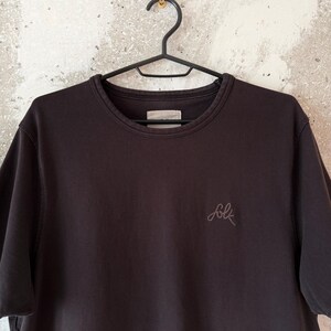 May include: A dark brown crew neck t-shirt hanging on a black hanger. The shirt has short sleeves and a small embroidered "folk" logo on the chest. The shirt is made of a soft, casual fabric.