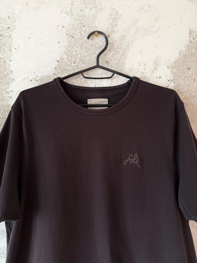 May include: A dark brown crew neck t-shirt hanging on a black hanger. The shirt has short sleeves and a small embroidered "folk" logo on the chest. The shirt is made of a soft, casual fabric.