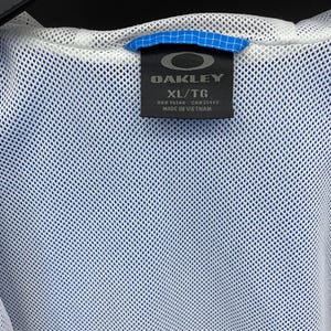 K&ouml;nnte beinhalten: Eine wei&szlig;e Mesh-Jacke mit einem blau-wei&szlig;en Etikett, auf dem "Oakley" und "XL/TG" steht. Das Etikett tr&auml;gt au&szlig;erdem die Aufschrift "RN# 98548 CA# 35460" und "Hergestellt in Vietnam."