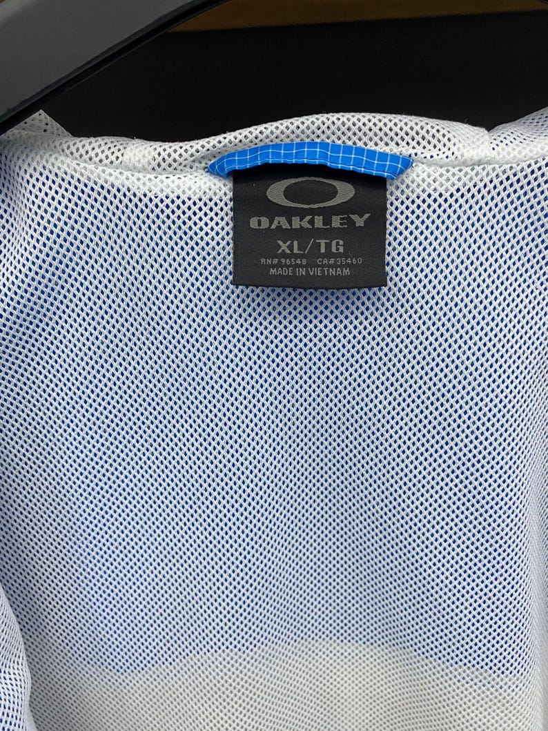 K&ouml;nnte beinhalten: Eine wei&szlig;e Mesh-Jacke mit einem blau-wei&szlig;en Etikett, auf dem "Oakley" und "XL/TG" steht. Das Etikett tr&auml;gt au&szlig;erdem die Aufschrift "RN# 98548 CA# 35460" und "Hergestellt in Vietnam."