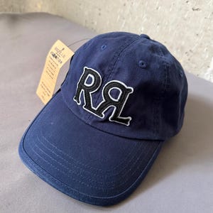 May include: A navy blue baseball cap featuring the embroidered letters RRL in black with a white outline on the front panel. The cap includes a curved brim, stitched eyelets, and a tan paper hang tag attached to the side with printed text.