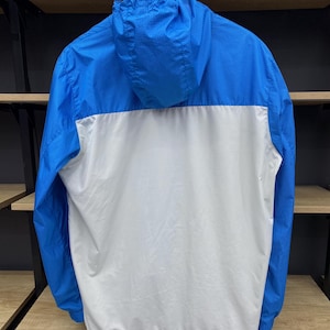 K&ouml;nnte beinhalten: Eine blaue und wei&szlig;e Windjacke mit Kapuze. Die Jacke hat ein kariertes Muster an den &Auml;rmeln und der Kapuze.