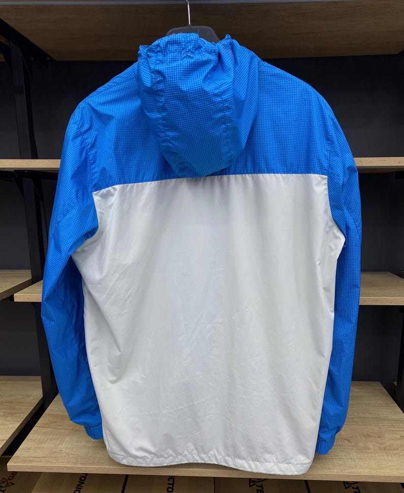 K&ouml;nnte beinhalten: Eine blaue und wei&szlig;e Windjacke mit Kapuze. Die Jacke hat ein kariertes Muster an den &Auml;rmeln und der Kapuze.