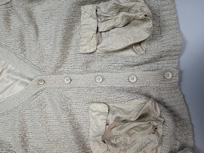 Op de afbeelding: Een beige gebreid vest met een V-hals, een knoopsluiting aan de voorzijde met vijf bijpassende ronde knopen en twee grote, gekreukte, lichtbeige stoffen zakken aan de onderkant. Het kledingstuk is voorzien van een gladde, lichtbeige voering.