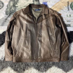 May include: A brown leather jacket with a zip-up front, two front pockets, and a collared neckline. The jacket has a worn, distressed appearance. The Polo Ralph Lauren logo is visible on the label.