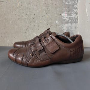 May include: A pair of brown leather sneakers with multiple hook-and-loop straps. The shoes have a rounded toe and decorative stitching. The sneakers are on a gray surface.