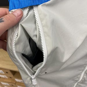 K&ouml;nnte beinhalten: Eine hellgraue Jacke mit Rei&szlig;verschluss. Der Rei&szlig;verschluss ist offen und zeigt eine schwarze Tasche im Inneren. Die Jacke hat ein blau-wei&szlig;es Karomuster am Kragen.