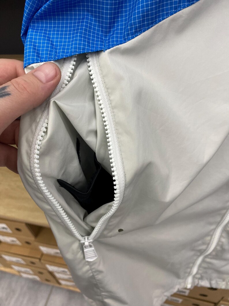 K&ouml;nnte beinhalten: Eine hellgraue Jacke mit Rei&szlig;verschluss. Der Rei&szlig;verschluss ist offen und zeigt eine schwarze Tasche im Inneren. Die Jacke hat ein blau-wei&szlig;es Karomuster am Kragen.
