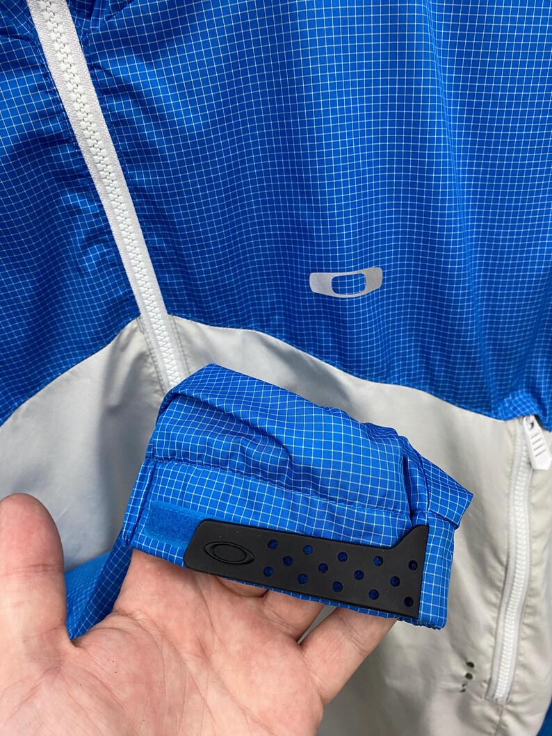 K&ouml;nnte beinhalten: Eine blaue und wei&szlig;e Windjacke mit Rei&szlig;verschluss und einem schwarzen Logo auf der Brust. Die Jacke hat ein Gittermuster und ein schwarzes Gummiband am &Auml;rmel.