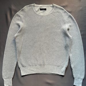 May include: A gray waffle knit sweater with a crew neck and long sleeves. The sweater has a small logo on the chest that says "AllSaints".