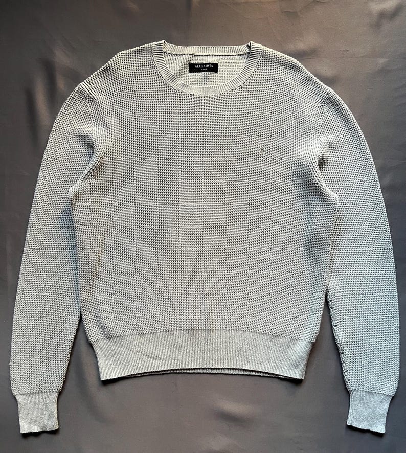 May include: A gray waffle knit sweater with a crew neck and long sleeves. The sweater has a small logo on the chest that says "AllSaints".
