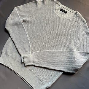 May include: A light gray waffle knit sweater with long sleeves and a crew neck. The sweater has a small black tag with the text "ALLSAINTS" on it.