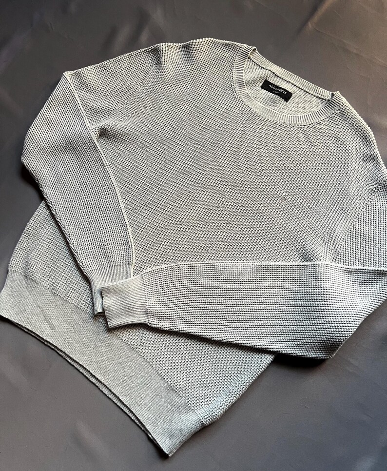 May include: A light gray waffle knit sweater with long sleeves and a crew neck. The sweater has a small black tag with the text "ALLSAINTS" on it.