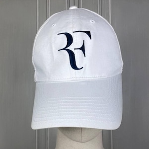 Può includere: Cappellino da baseball bianco con visiera curva. Il cappellino presenta un logo "RF" blu navy ricamato sul davanti. Il cappello è realizzato in un tessuto liscio e ha un bottone sulla parte superiore. Il cappellino è esposto su una testa di manichino.