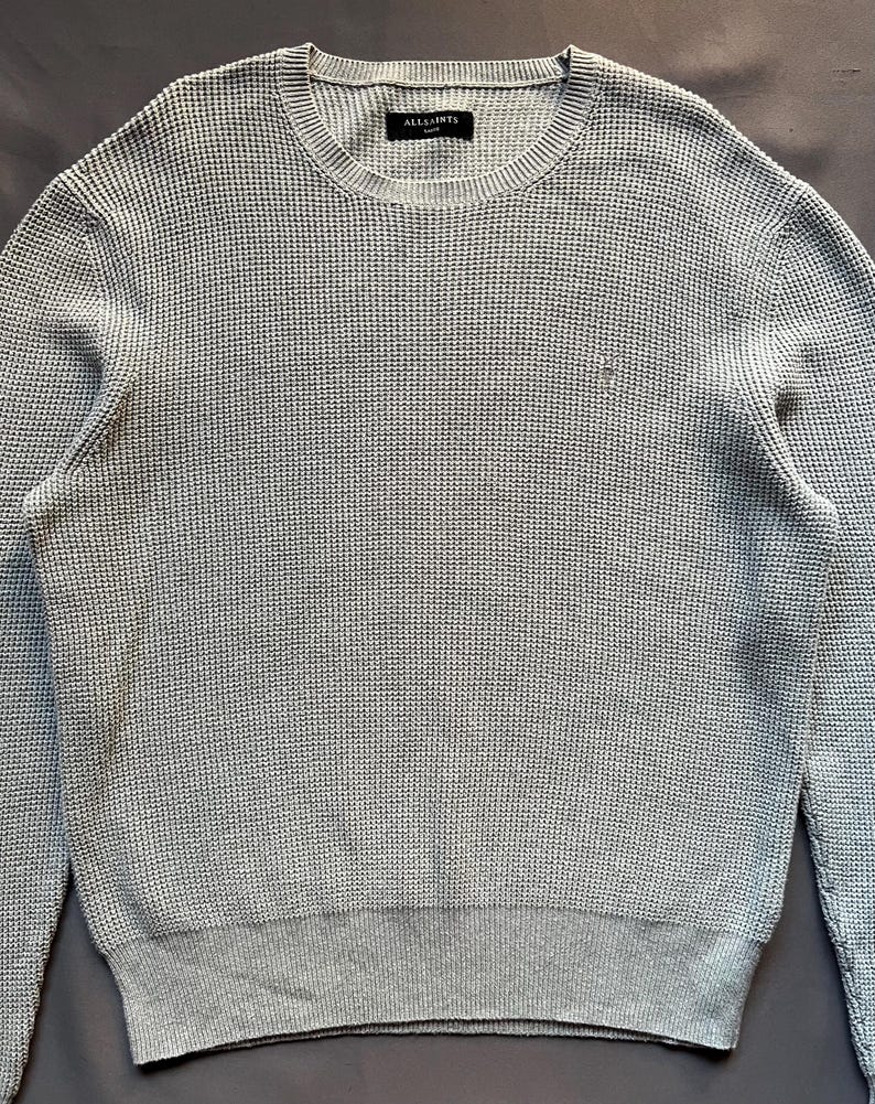 May include: A light gray waffle knit sweater with a small logo on the left chest. The sweater has a crew neck and long sleeves.
